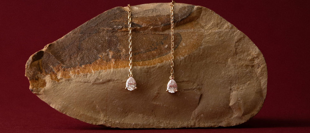 Anate's Talya pear diamond drop earrings in solid yellow gold suspended over a fossil stone, timeless craftsmanship